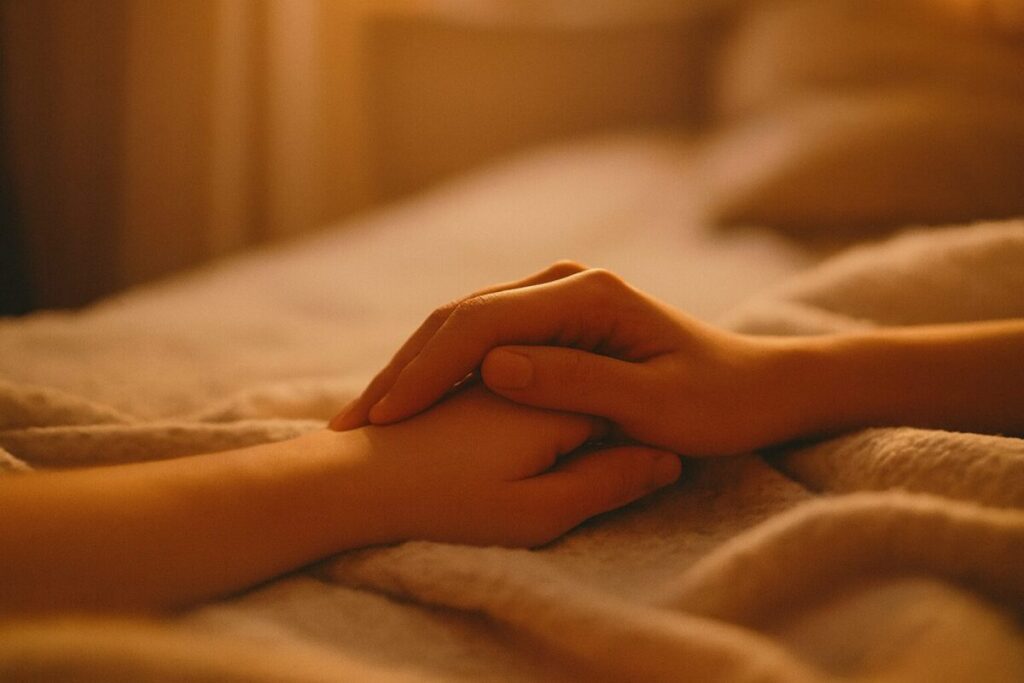 Horizontal close-up photograph of two hands resting gently on a soft beige blanket. One hand lightly holds the other in a self-soothing gesture, bathed in warm evening light. The background is softly blurred, suggesting a peaceful bedroom. The mood is intimate, calm, and emotionally tender, symbolizing self-care and quiet reassurance.